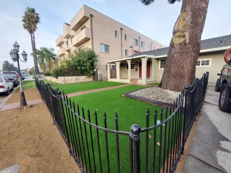 Artificial turf lawn behind a black iron fence beside a palm tree and apartment building